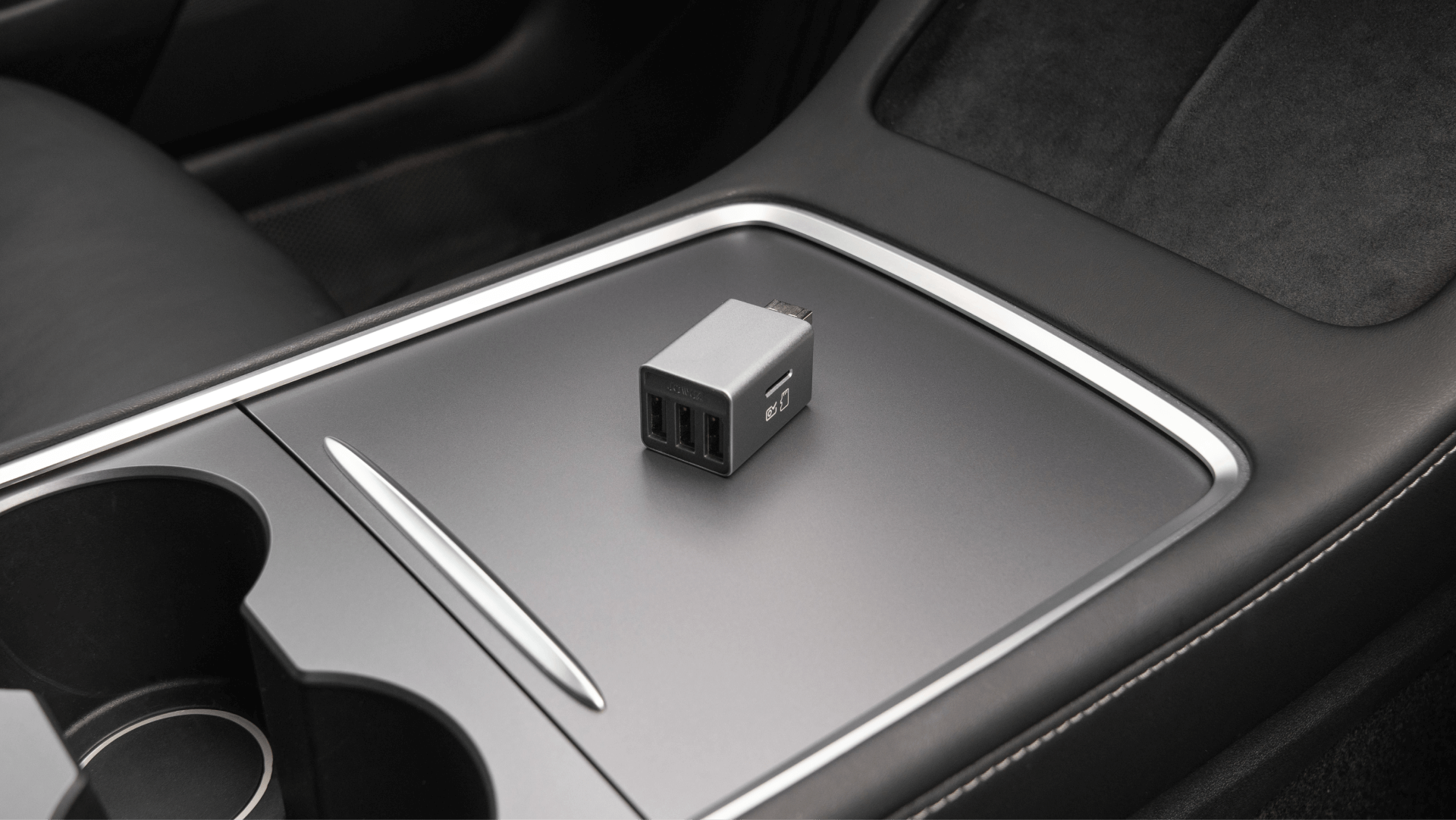 A USB car charger with two ports sits on the stylish JOWUA 3 Port USB HUB with DashCam Reader within the vehicle's interior, adjacent to the cup holders. The console boasts a modern design, featuring a smooth and dark surface.