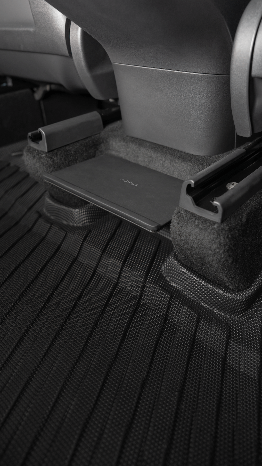 The image shows the interior of a Tesla Model Y, highlighting the rear seat area. On the carpeted floor, there's a rectangular object, which could be part of the JOWUA Model Y Tissue Box Holder Set. Also visible are the lower part of a car seat and a metal rail system.