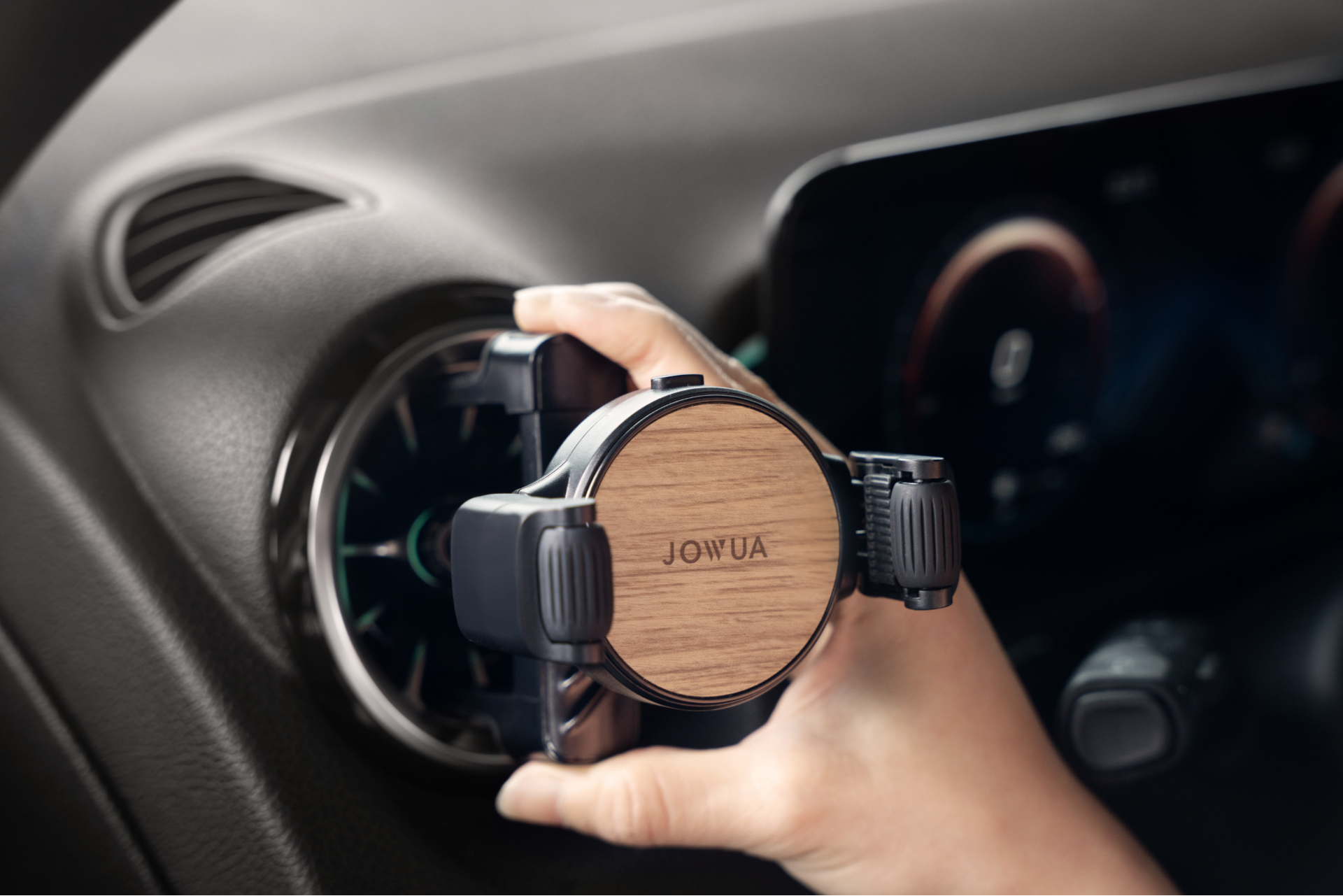 A person is installing a JOWUA Universal Air Vent Car Mount onto a car's dash. This mount features a wood finish, offers 360-degree rotation, and comes equipped with an adjustable clamp to keep your device secure. In the background, part of the dashboard screen can be seen.