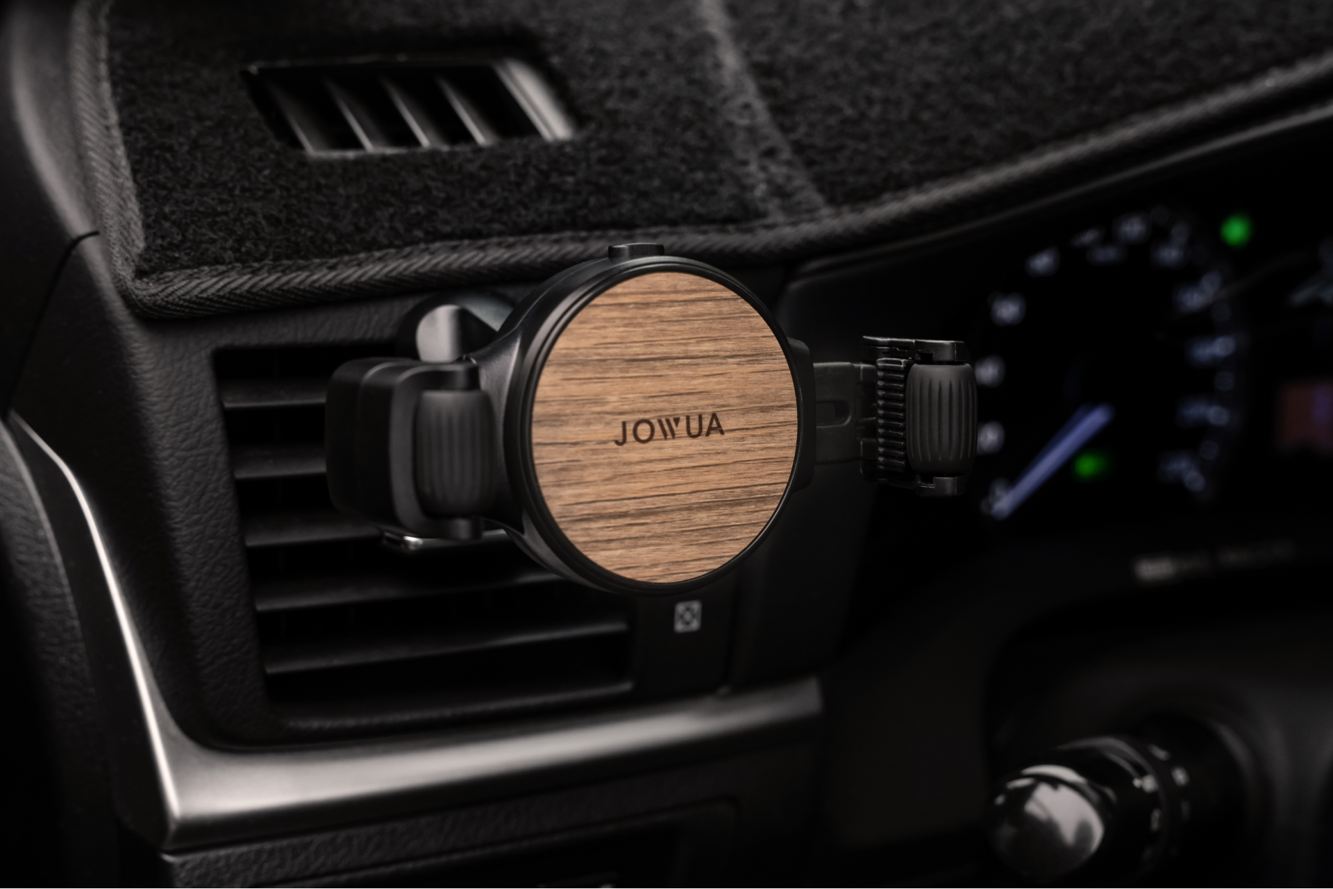 The JOWUA Universal Air Vent Car Mount, featuring a wooden finish and adjustable clamps, is attached to the car dashboard's air vent. The interior showcases black upholstery, with a blurred background highlighting the speedometer and other controls.