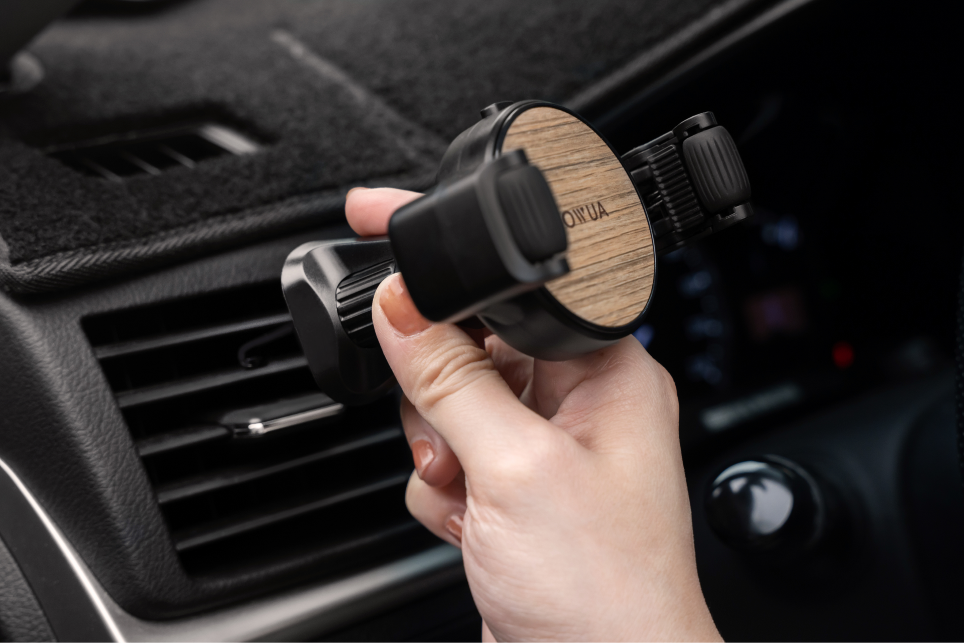 A person holds the JOWUA Universal Air Vent Car Mount featuring a sleek wooden finish and adjustable clamps, preparing to install it onto a vehicle's air vent. The dashboard and control panel are visible in the background, set for convenient 360-degree rotation.